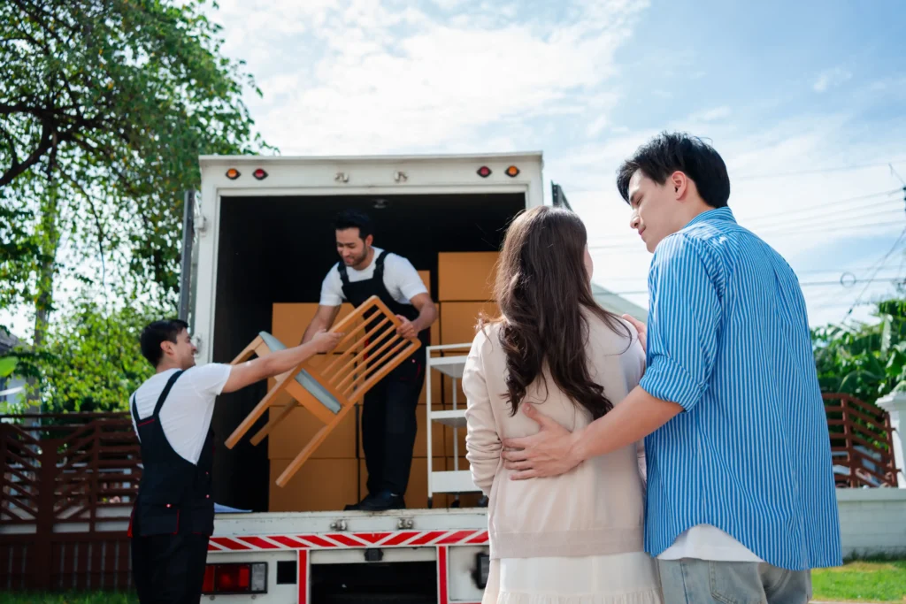 A couple embraces while observing movers unloading furniture from a truck on a sunny day, conveying a sense of new beginnings and excitement.