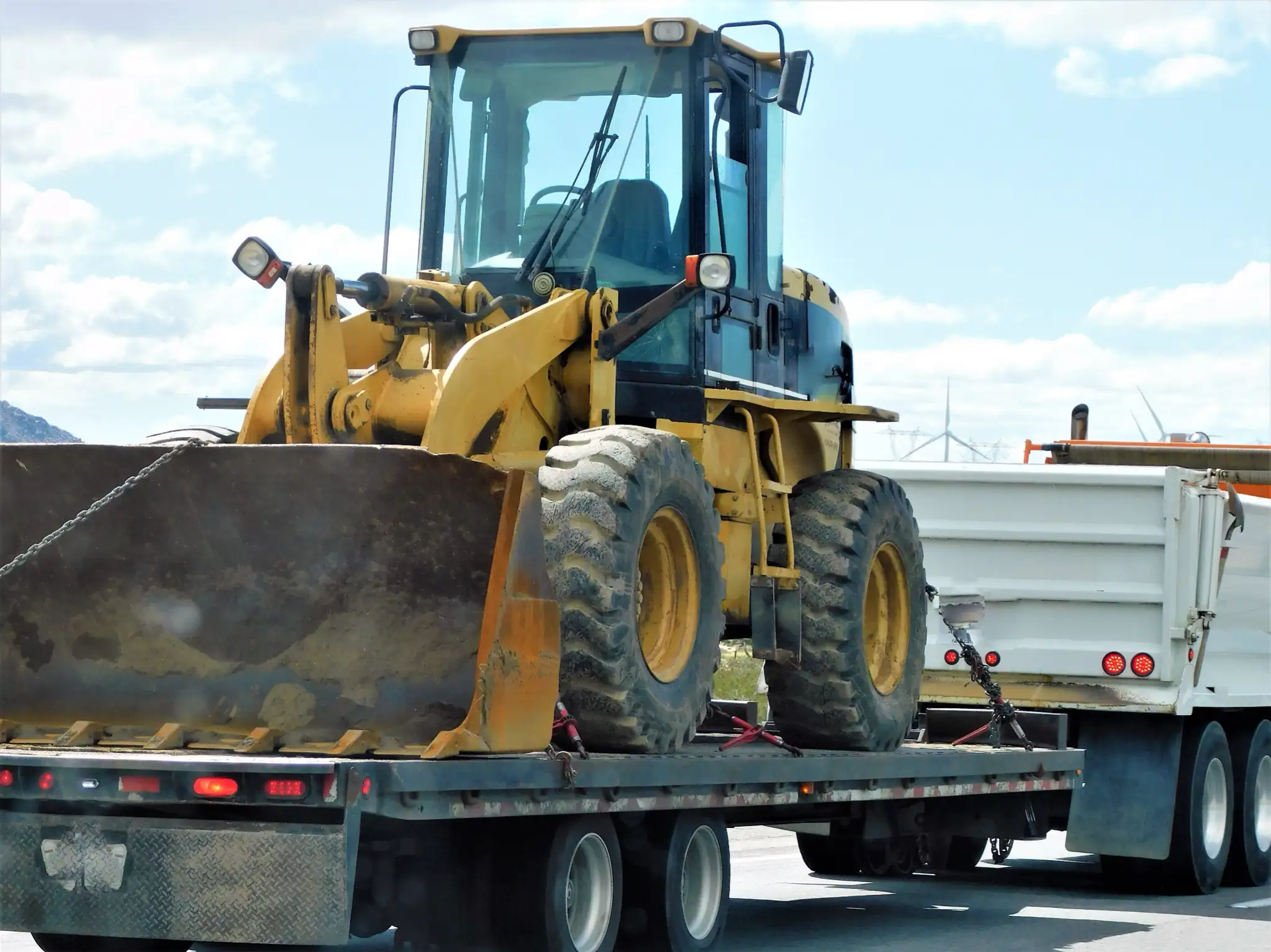 Yellow front loader secured on a flatbed trailer, part of a large truck convoy. The scene is sunny, depicting industrial transport on a highway.