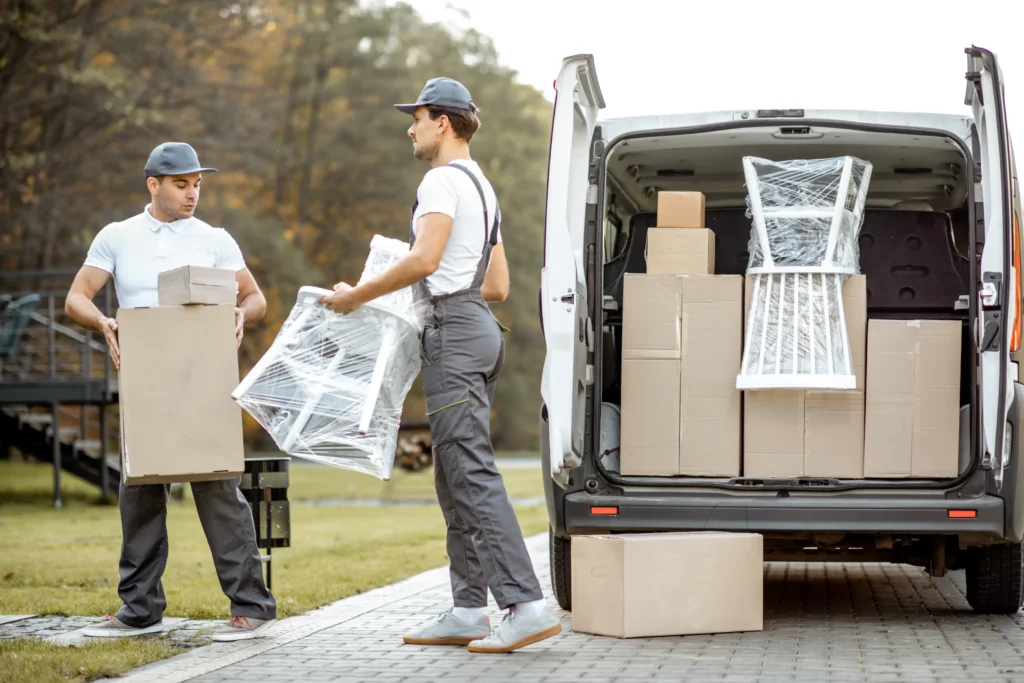 Two movers unload a white van on a sunny day. One carries a wrapped chair while the other holds boxes. The background shows a grassy park.