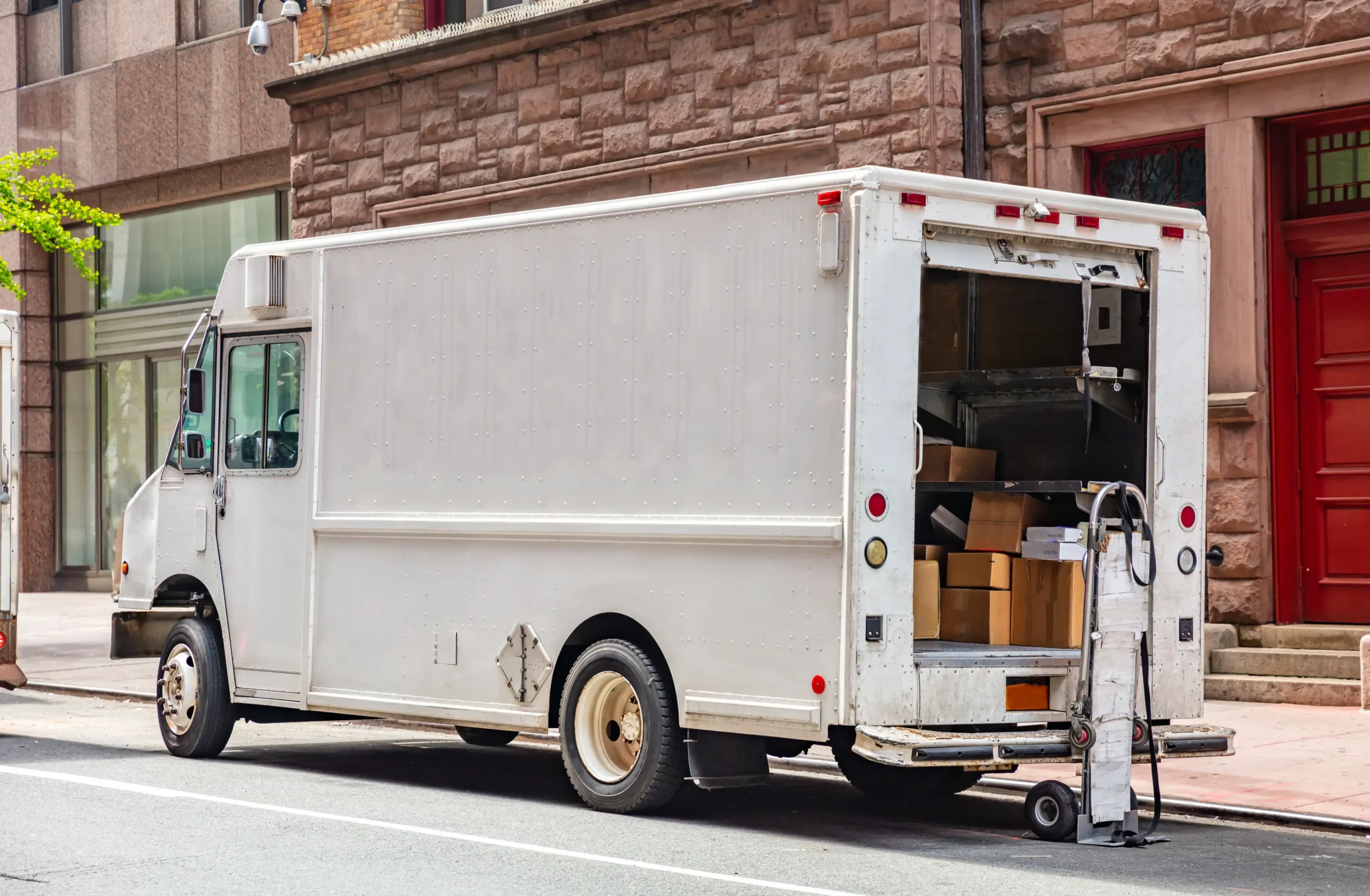 White delivery truck parked on city street with open back door, loaded with various cardboard boxes. A dolly leans against the truck.